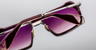 Close-up of Jacques Marie Mage GT-R sunglasses featuring a geometric frame, maroon rims, gold accents, and purple gradient lenses set against a plain light gray background.