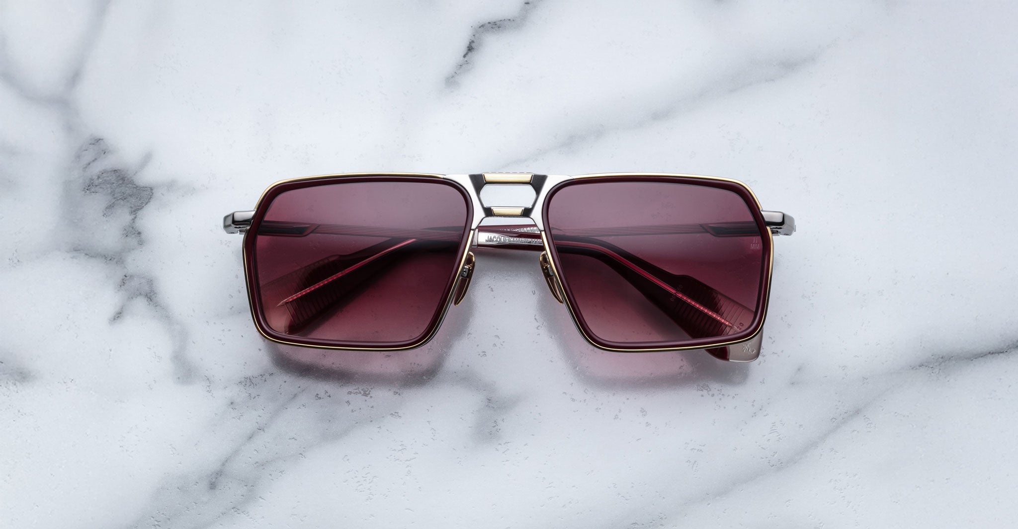 A pair of Jacques Marie Mage GT-R rectangular sunglasses with silver frames and dark red lenses is placed on a white marble surface.
