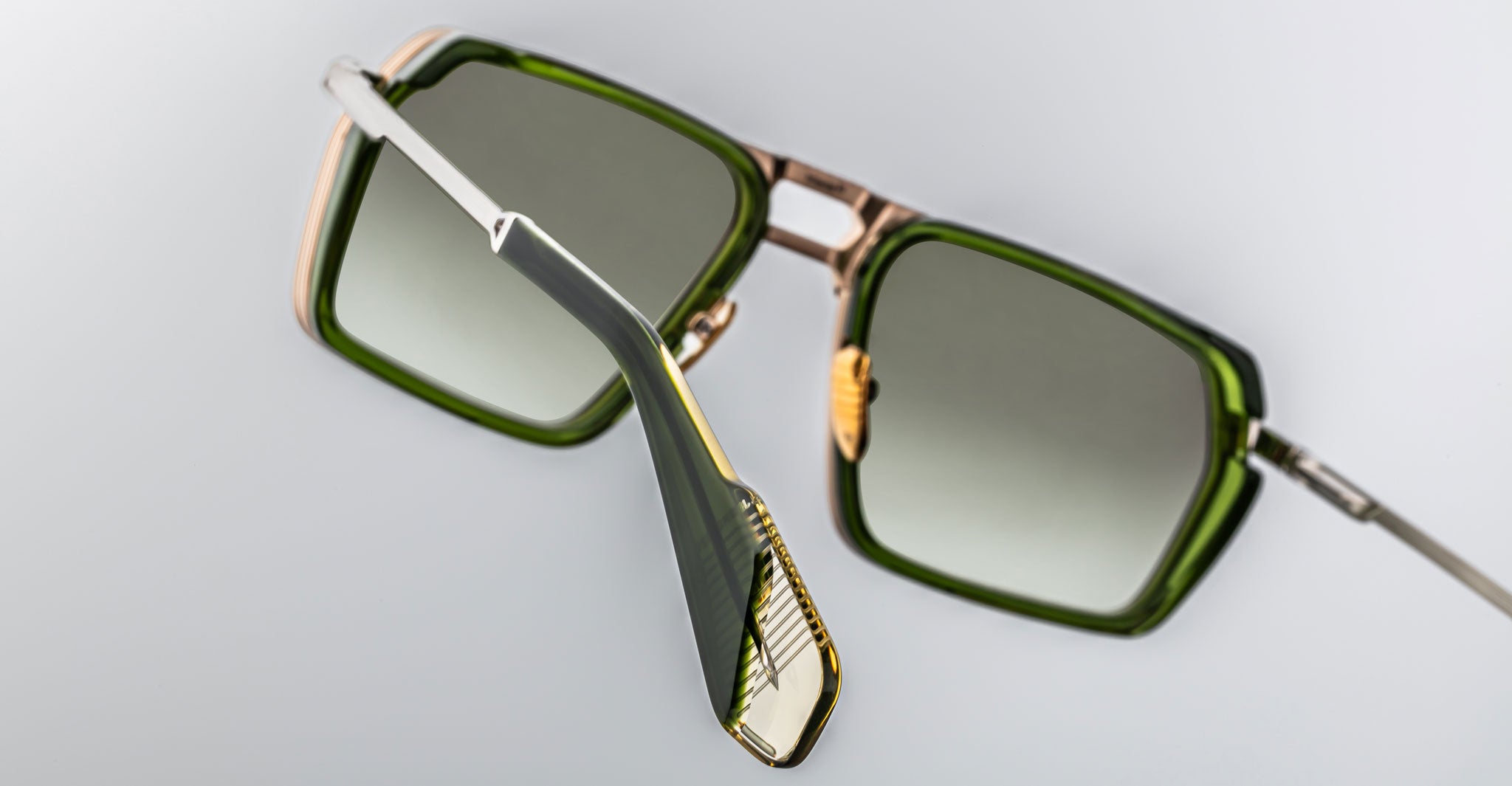 Close-up of Jacques Marie Mage GT-R sunglasses featuring square green-tinted lenses, gold accents, and black-and-gold detailed temple arms against a plain light background.