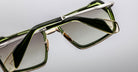 Close-up of Jacques Marie Mage GT-R sunglasses, featuring green rectangular frames with gold and silver accents, folded on a light grey surface. The lenses display a stylish gradient tint.