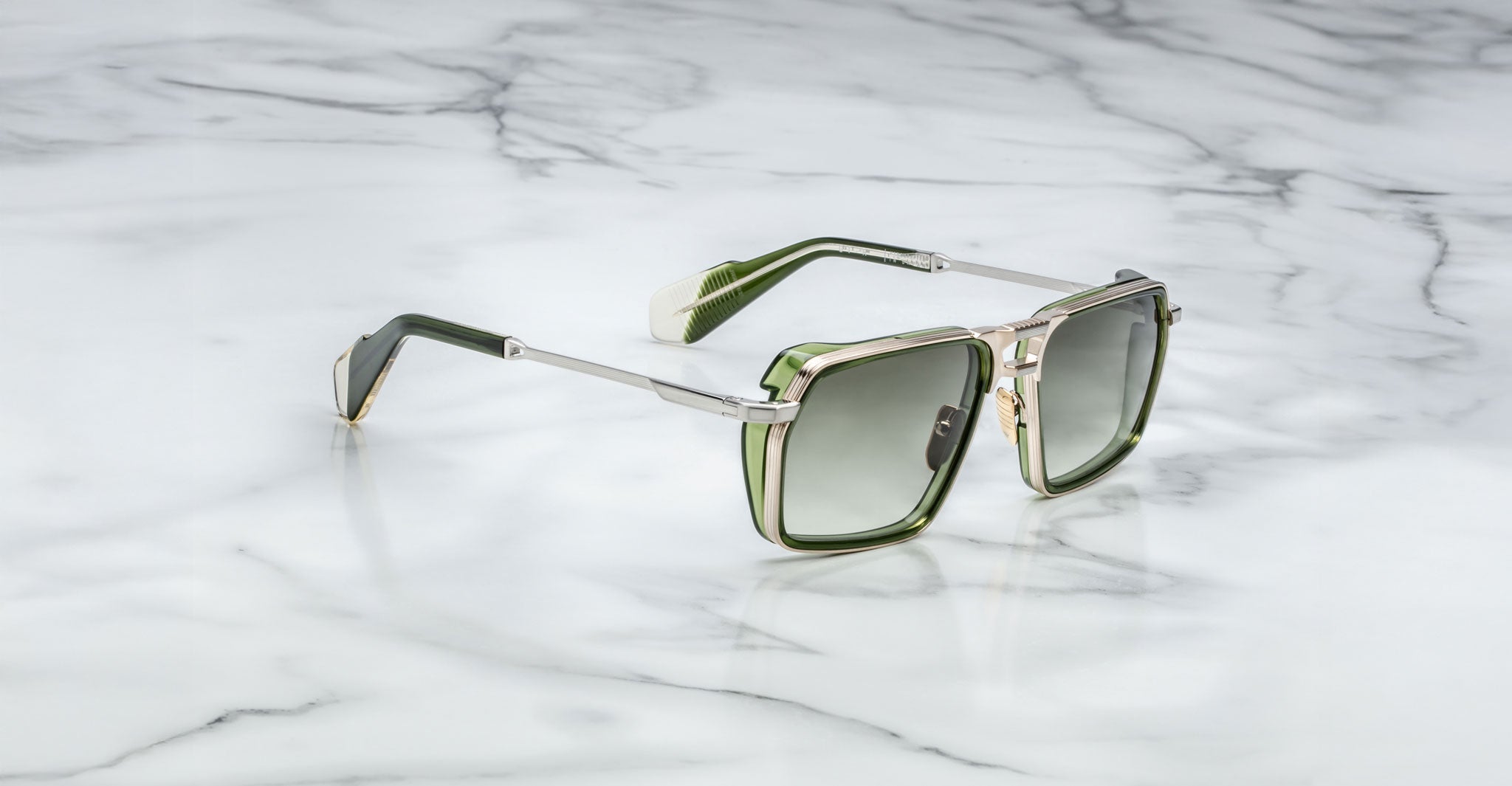 A pair of Jacques Marie Mage GT-R green and silver square-framed sunglasses with dark lenses rests on a white marble surface.