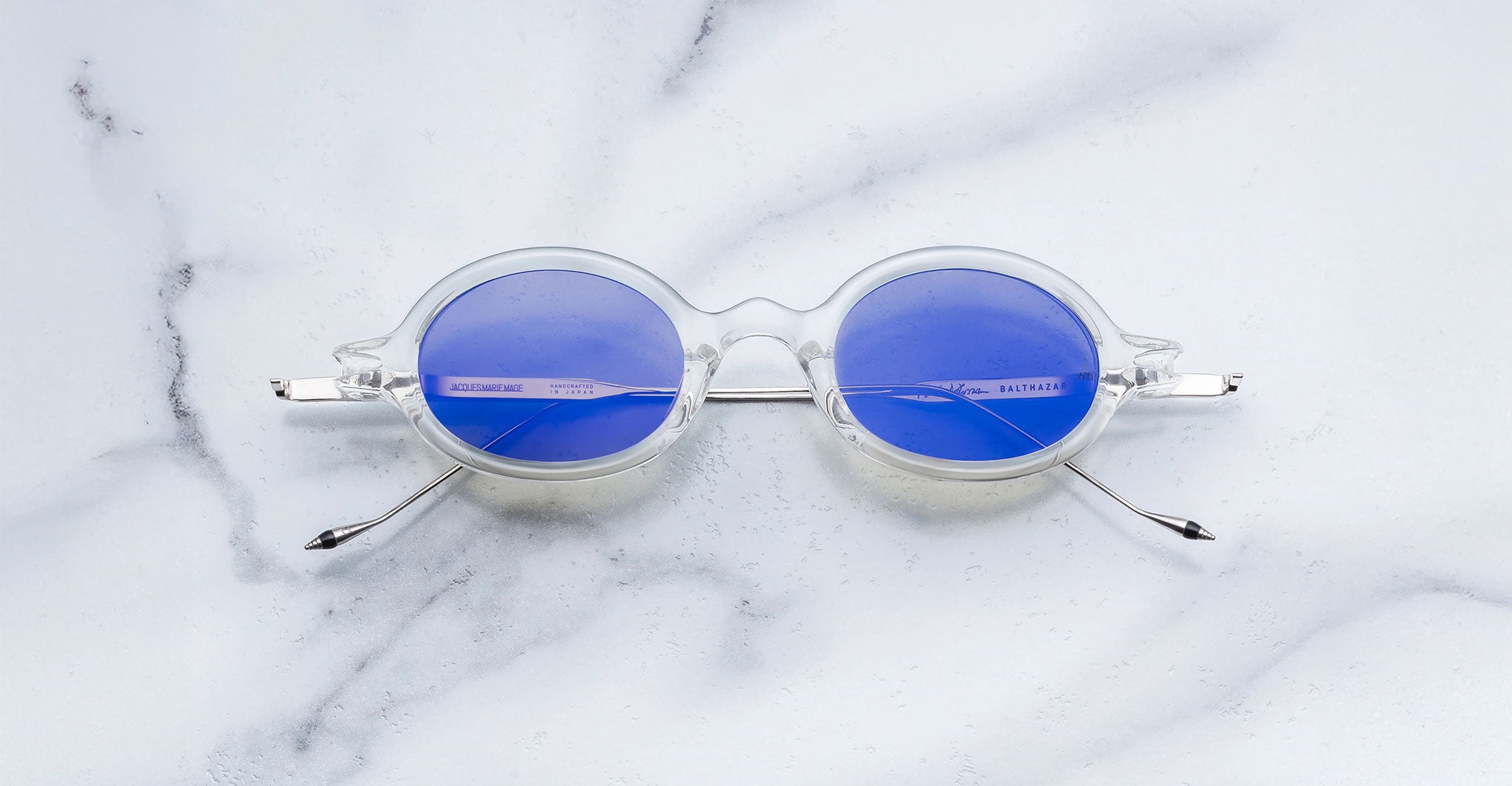 Jacques Marie Mage Balthazar round clear sunglasses with blue lenses, top view, eyewear displayed on surface.