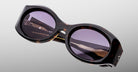 Close-up of Jacques Marie Mage Gia sunglasses—large, round, dark tortoiseshell acetate frames with purple-tinted lenses—handcrafted in Japan and reflecting a modern striped architectural structure.
