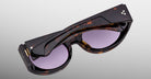The Jacques Marie Mage Gia sunglasses, handcrafted in Japan with an oval acetate frame and purple lenses, are neatly folded and displayed against a plain grey background.