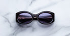 The Jacques Marie Mage Gia, oval acetate sunglasses by Jacques Marie Mage, handcrafted in Japan with purple-tinted lenses, rest on a white marble surface.
