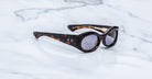 A pair of Jacques Marie Mage Gia oval acetate sunglasses by Jacques Marie Mage, handcrafted in Japan with tortoiseshell frames and round dark lenses, displayed on a white marble surface.