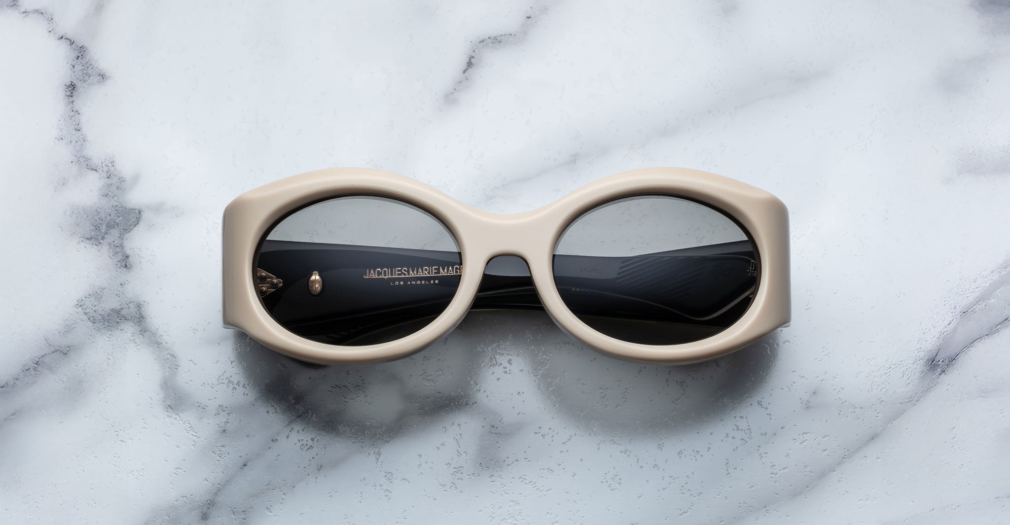 A pair of Jacques Marie Mage Gia beige, thick-framed sunglasses with dark lenses rests on a white marble surface with grey veins, with faint brand details visible on the inside of the left lens.