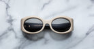 A pair of Jacques Marie Mage Gia beige, thick-framed sunglasses with dark lenses rests on a white marble surface with grey veins, with faint brand details visible on the inside of the left lens.