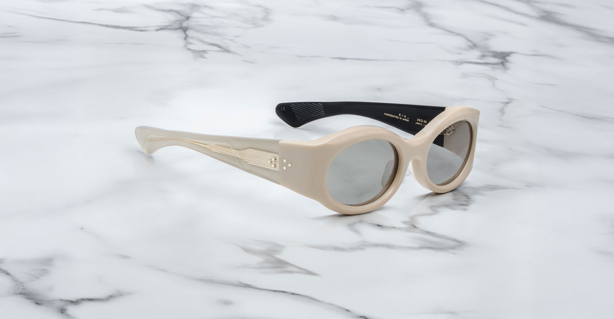 A pair of Jacques Marie Mage Gia beige, thick-framed sunglasses with dark lenses sits on a white marble surface with grey veins, with faint brand details visible on the inside of the left lens.