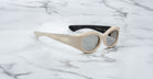 A pair of Jacques Marie Mage Gia beige, thick-framed sunglasses with dark lenses sits on a white marble surface with grey veins, with faint brand details visible on the inside of the left lens.