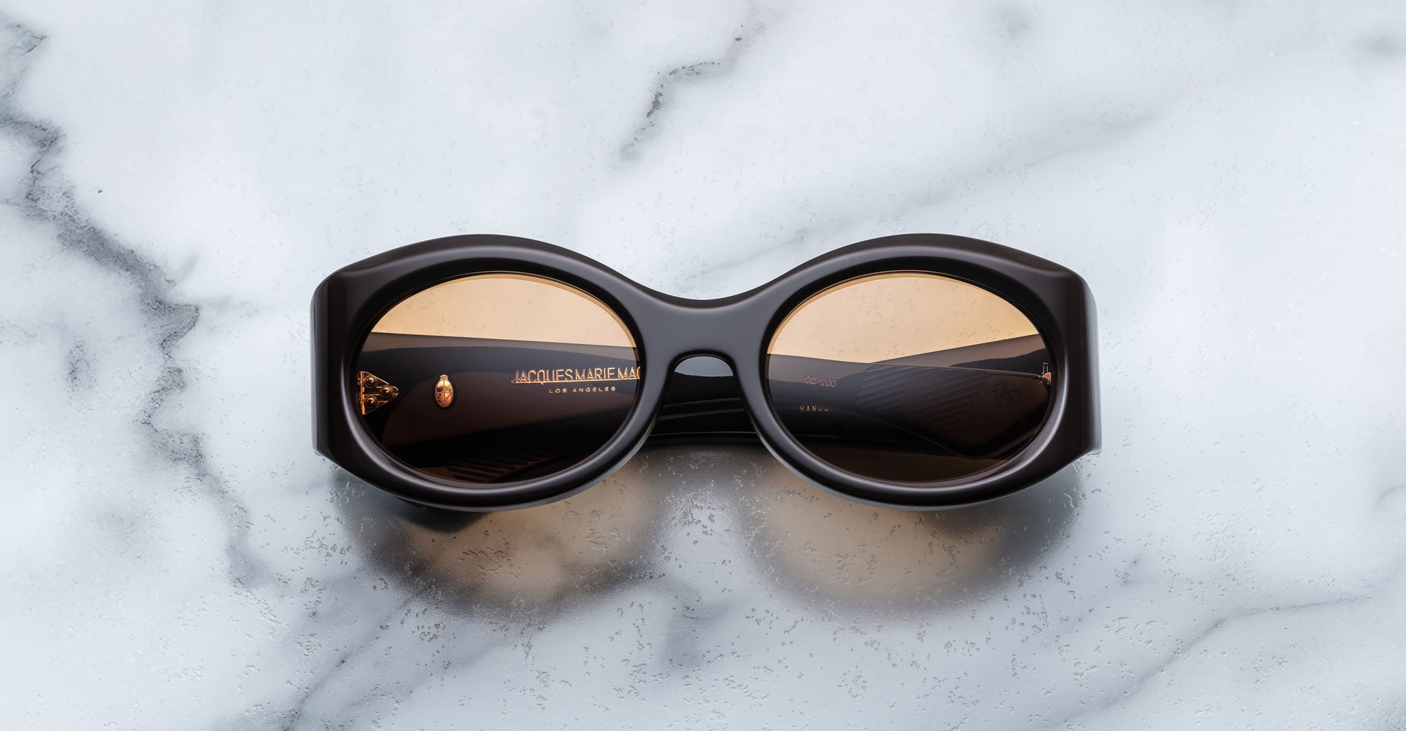 A pair of Jacques Marie Mage Gia walnut, thick-framed sunglasses with orange lenses rests on a white marble surface with grey veins, with faint brand details visible on the inside of the left lens.