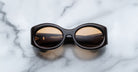 A pair of Jacques Marie Mage Gia walnut, thick-framed sunglasses with orange lenses rests on a white marble surface with grey veins, with faint brand details visible on the inside of the left lens.