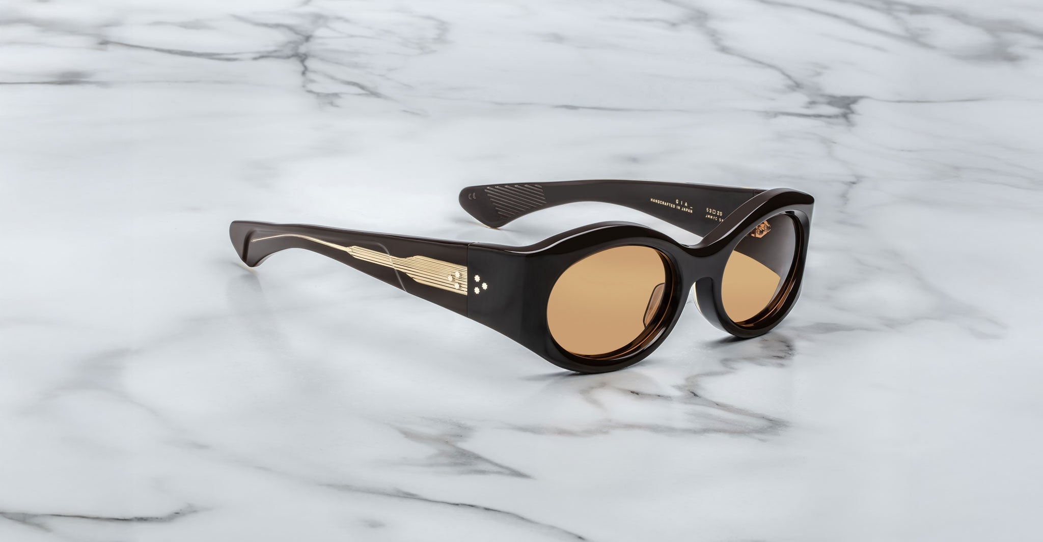 A pair of Jacques Marie Mage Gia walnut color, thick-framed sunglasses with orange lenses sits on a white marble surface with grey veins, with faint brand details visible on the inside of the left lens.
