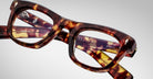 Close-up of Jacques Marie Mage Godard tortoiseshell eyeglasses with thick rectangular frames, resting on their side. Product details are visible on the inner arms against a light gray background.