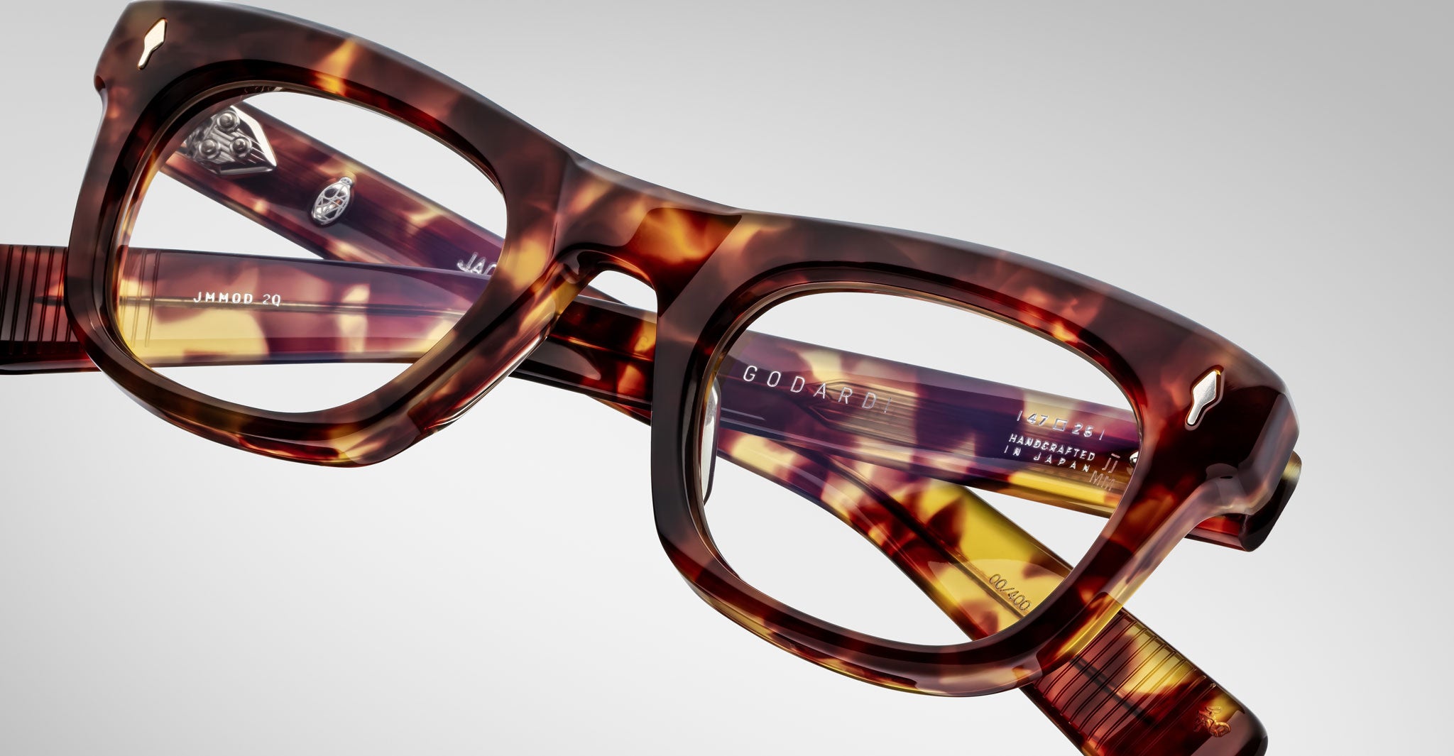 Close-up of Jacques Marie Mage Godard tortoiseshell eyeglasses with thick rectangular frames, resting on their side. Product details are visible on the inner arms against a light gray background.