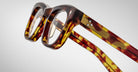 Close-up of Jacques Marie Mage Godard eyeglasses featuring thick, brown and yellow tortoiseshell-patterned plastic frames, shown at an angle highlighting the front and side arms against a plain light background.