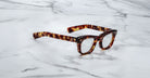 A pair of Jacques Marie Mage Godard thick-rimmed, tortoiseshell eyeglasses rests on a white marble surface with gray veining.
