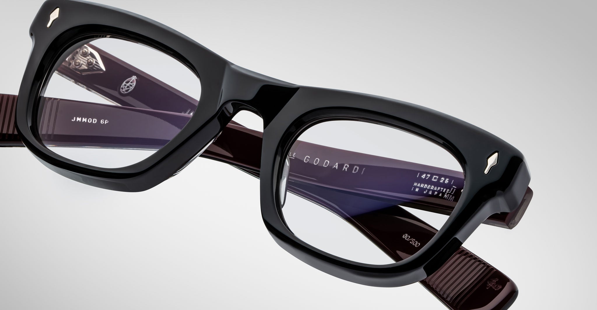Close-up view of Jacques Marie Mage Godard black thick-rimmed eyeglasses with clear lenses. The inside arms show brand markings and model details. Set against a plain, light-colored background.