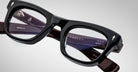 Close-up view of Jacques Marie Mage Godard black thick-rimmed eyeglasses with clear lenses. The inside arms show brand markings and model details. Set against a plain, light-colored background.