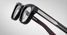 A close-up of Jacques Marie Mage Godard thick-rimmed black eyeglasses with silver detailing along the temples, resting on a neutral gray background. White text is inscribed inside the arm.