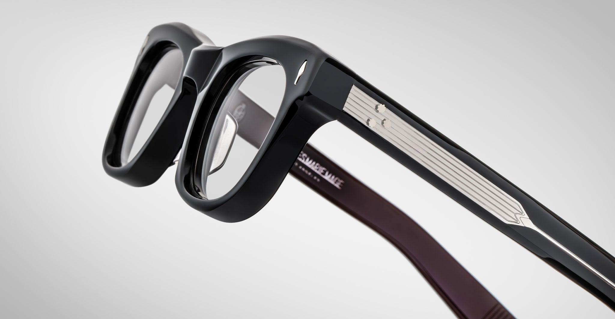 A close-up of Jacques Marie Mage Godard thick-rimmed black eyeglasses with silver detailing along the temples, resting on a neutral gray background. White text is inscribed inside the arm.