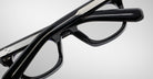 Close-up of Jacques Marie Mage Godard black eyeglasses with silver-detailed temples, folded against a plain light gray background.