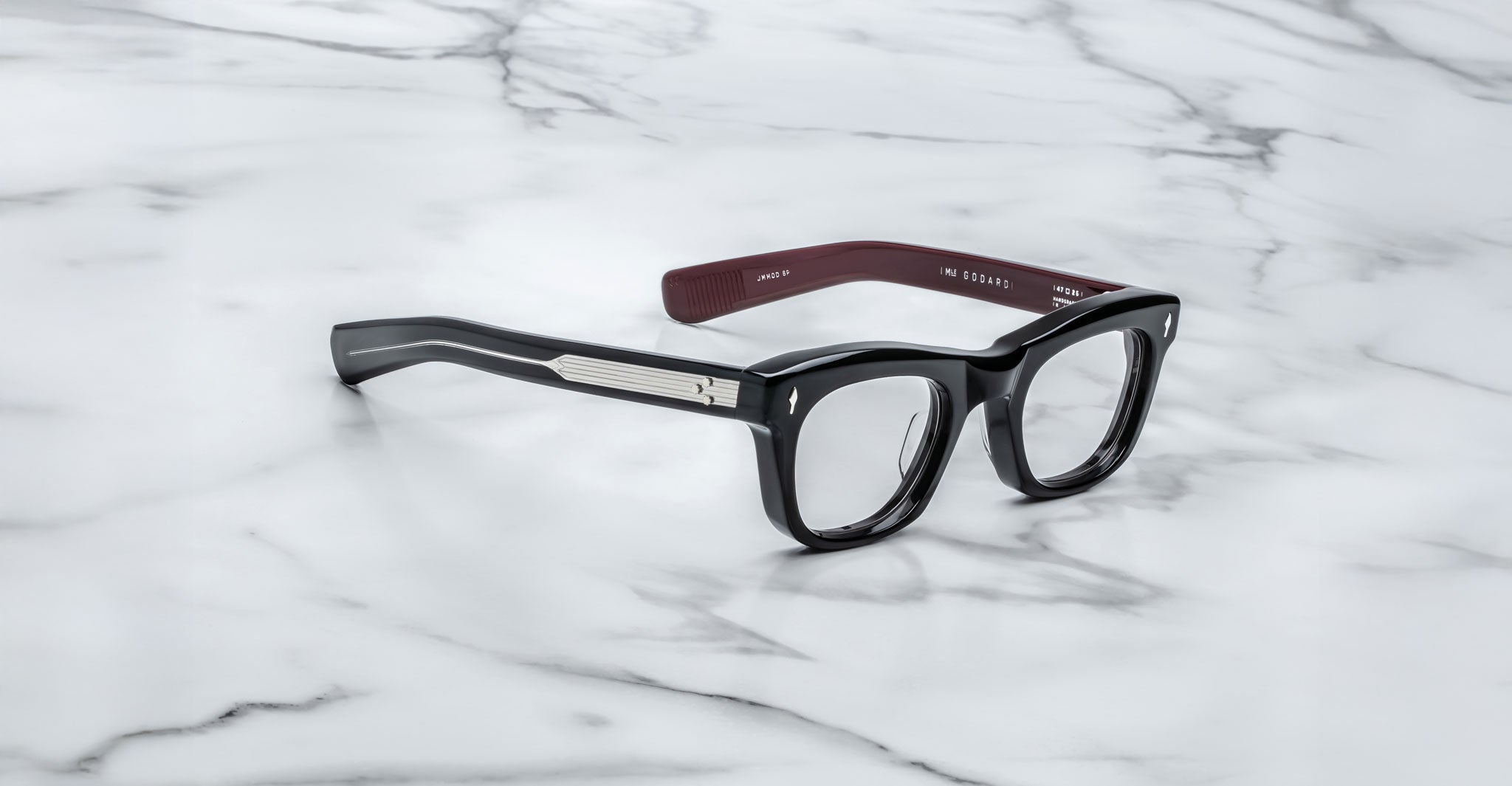 A pair of Jacques Marie Mage Godard black rectangular eyeglasses with thick frames, silver side accents, and brown temple interiors rests on a white marble surface.
