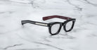 A pair of Jacques Marie Mage Godard black rectangular eyeglasses with thick frames, silver side accents, and brown temple interiors rests on a white marble surface.