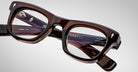 A close-up of rectangular, thick-framed Jacques Marie Mage Godard brown eyeglasses with translucent lenses on a plain gray background. The brand name and model details appear on the inside of the frame arms.