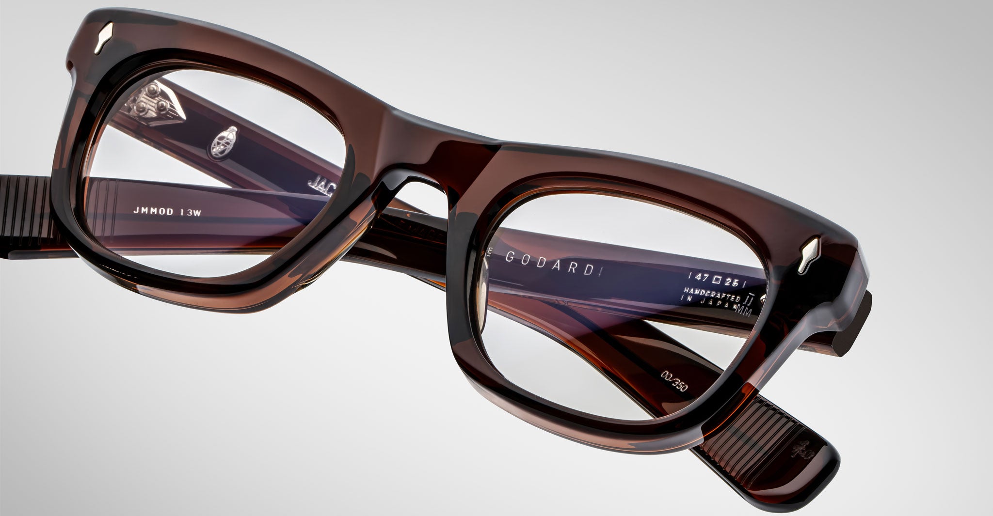 A close-up of rectangular, thick-framed Jacques Marie Mage Godard brown eyeglasses with translucent lenses on a plain gray background. The brand name and model details appear on the inside of the frame arms.