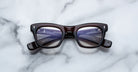 A pair of Jacques Marie Mage Godard dark-rimmed rectangular eyeglasses with clear lenses rests on a white marble surface, showing the brand name and specifications on the inside of the temple arms.