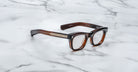 The Jacques Marie Mage Godard eyeglasses feature thick brown tortoiseshell rectangular frames with gold detailing on the temples, displayed on a white marble surface.