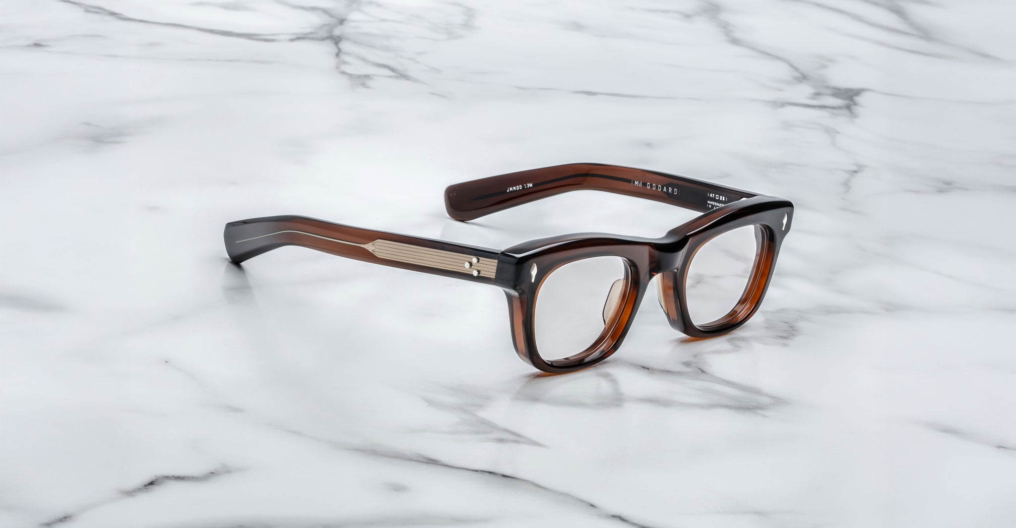 The Jacques Marie Mage Godard eyeglasses feature thick brown tortoiseshell rectangular frames with gold detailing on the temples, displayed on a white marble surface.