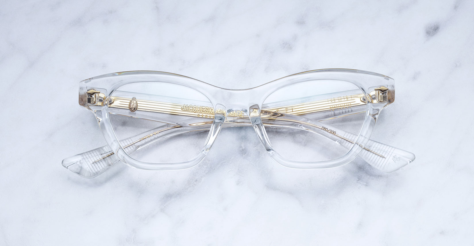 Jacques Marie Mage Grace clear cat-eye eyeglasses with gold accents, displayed on a white marble surface. Viewed from above, the arms are folded neatly behind the lenses.