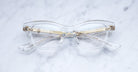Jacques Marie Mage Grace clear cat-eye eyeglasses with gold accents, displayed on a white marble surface. Viewed from above, the arms are folded neatly behind the lenses.