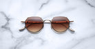 The Jacques Marie Mage Hartana features round, gold frames and brown gradient lenses, shown resting on a white marble surface with gray veining.