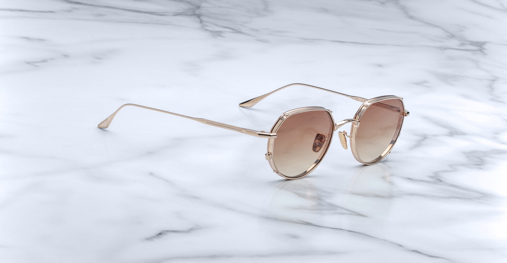 The Jacques Marie Mage Hartana sunglasses feature round, gold frames with light brown tinted lenses and slim metallic temples, displayed on a white marble surface with grey veining.