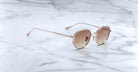 The Jacques Marie Mage Hartana sunglasses feature round, gold frames with light brown tinted lenses and slim metallic temples, displayed on a white marble surface with grey veining.