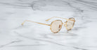 Jacques Marie Mage Hartana gold-framed round sunglasses with lightly tinted lenses rest on a smooth white marble surface with gray veining.