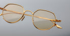 Close-up of Jacques Marie Mage Hartana gold-framed, round eyeglasses with light tinted lenses on a plain light gray background, showcasing fine detailing and visible inscriptions on the arms.