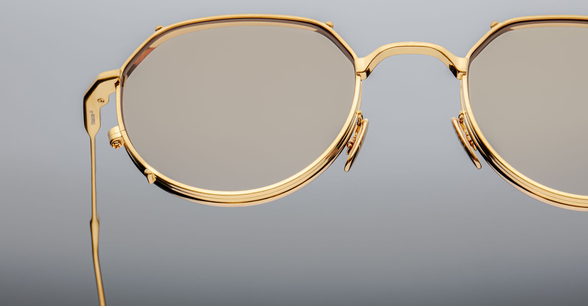 Close-up of Jacques Marie Mage Hartana eyeglasses with stylish gold frames and round lenses, set against a plain gray background. Only the right half of the glasses is visible in the image.