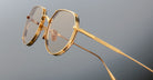 Close-up of Jacques Marie Mage Hartana eyeglasses featuring stylish gold frames, round lenses with a light brown tint, and thin temples, set against a plain grey background.