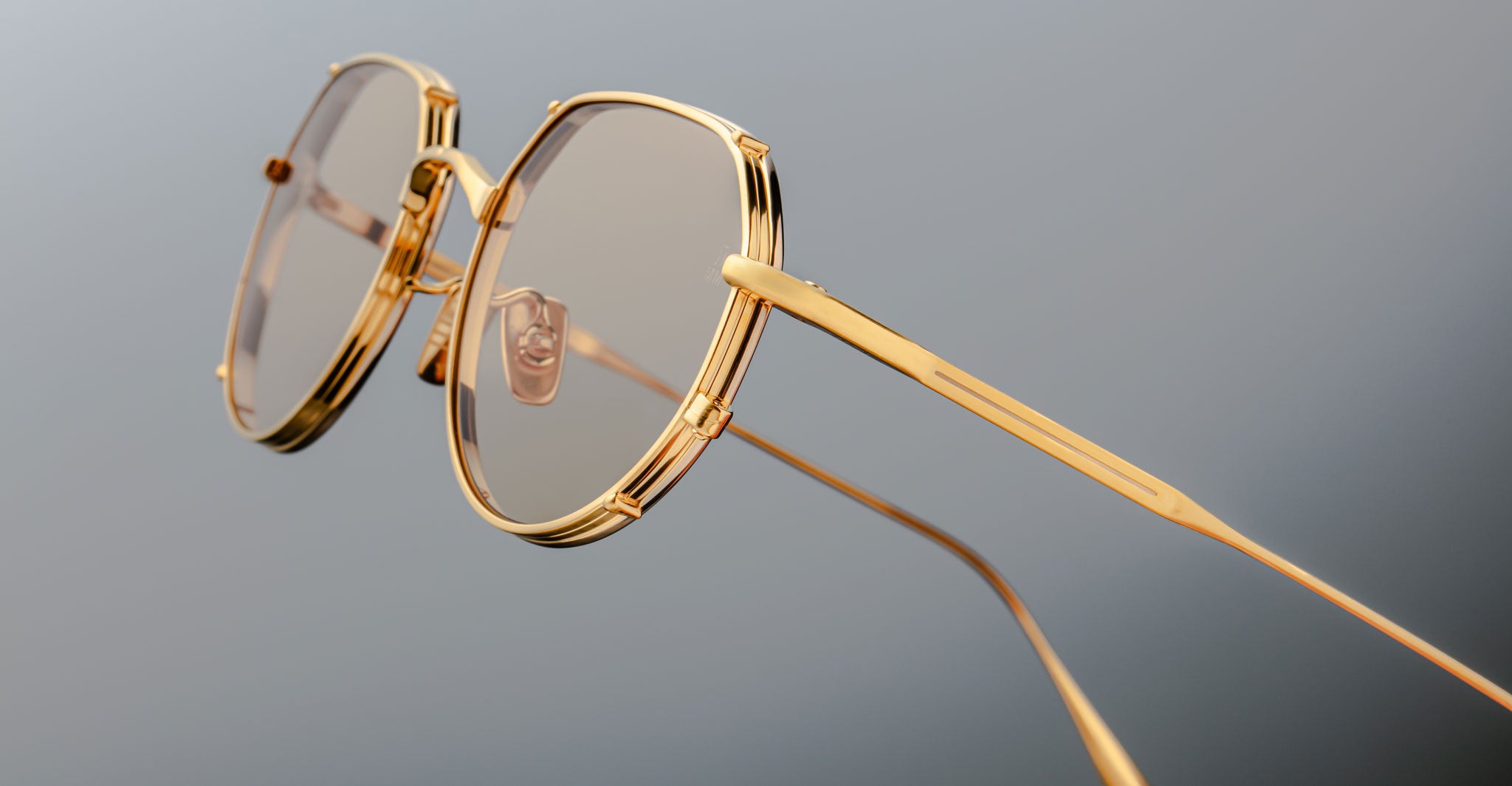 Close-up of Jacques Marie Mage Hartana eyeglasses featuring stylish gold frames, round lenses with a light brown tint, and thin temples, set against a plain grey background.