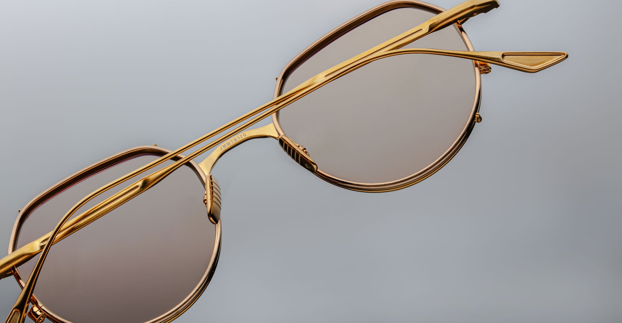 Close-up of Jacques Marie Mage Hartana sunglasses with stylish gold frames and brown tinted lenses, displayed on a plain gray background. The image showcases the sleek metal frame and modern design details.