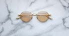 The Jacques Marie Mage Hartana features gold frames and round brown-tinted lenses, displayed on a white marble surface with grey veining.