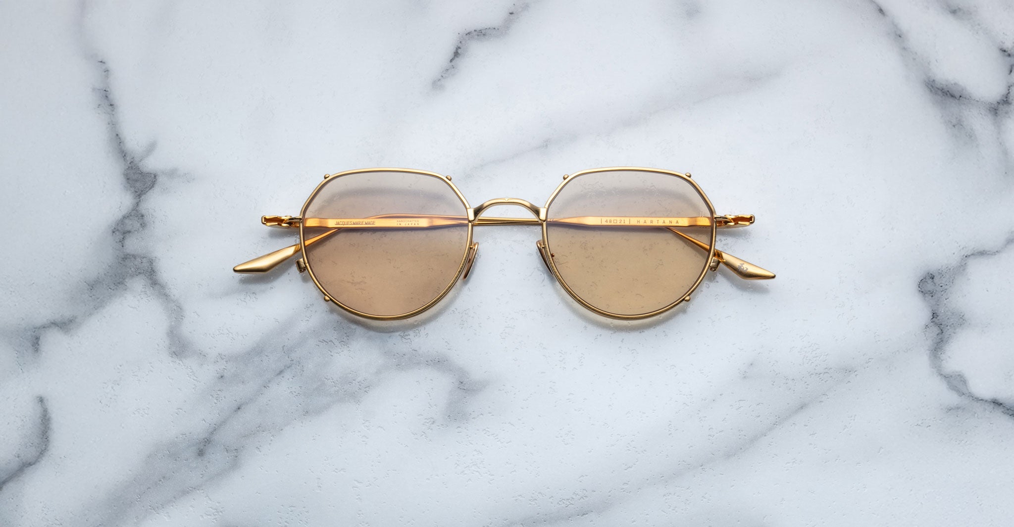 The Jacques Marie Mage Hartana features gold frames and round brown-tinted lenses, displayed on a white marble surface with grey veining.