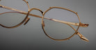 Close-up of Jacques Marie Mage Hartana gold metal eyeglass frames with clear lenses, detailed hinges, and engraved text on the arms, set against a neutral gray background.
