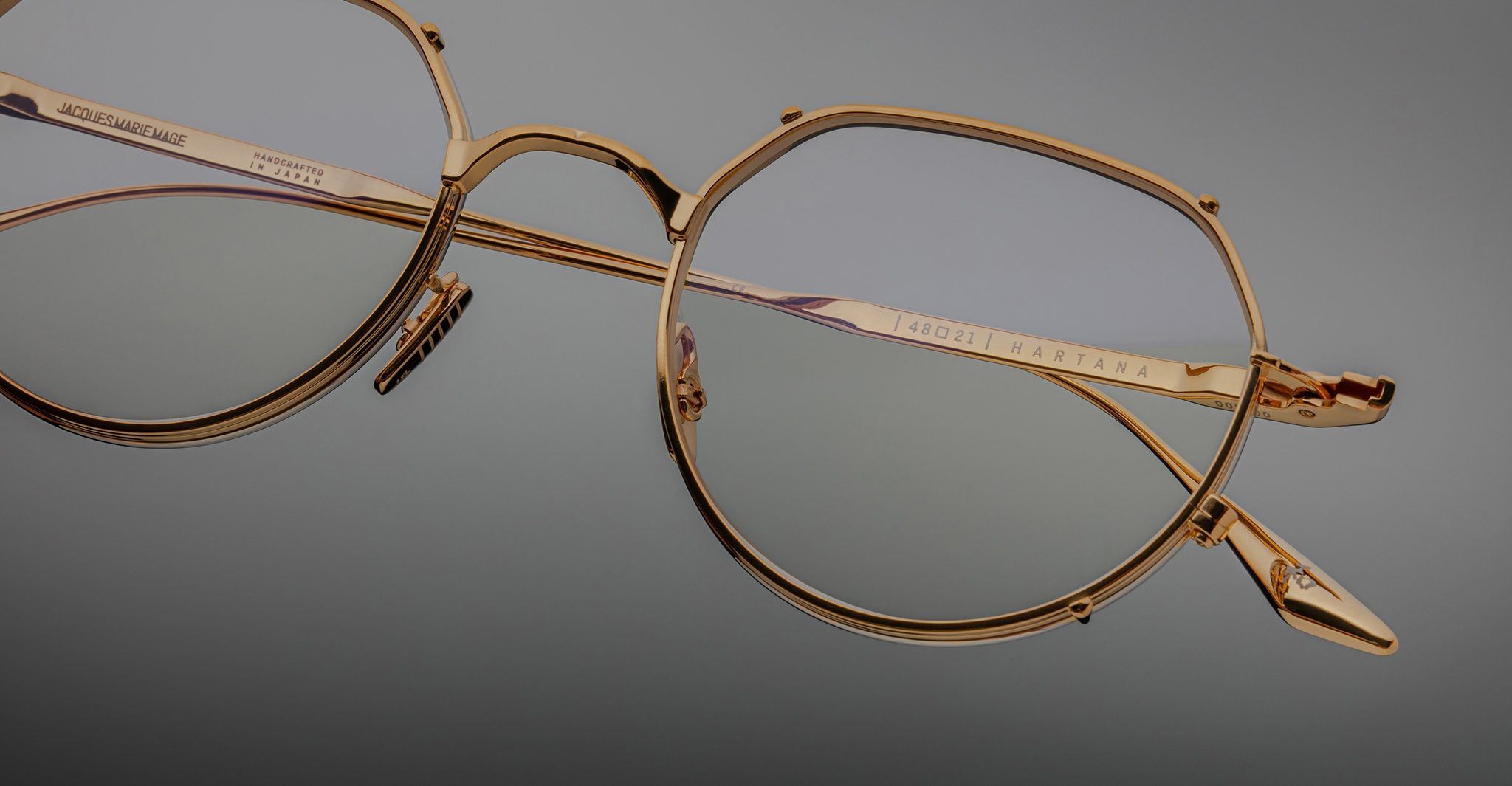 Close-up of Jacques Marie Mage Hartana gold metal eyeglass frames with clear lenses, detailed hinges, and engraved text on the arms, set against a neutral gray background.