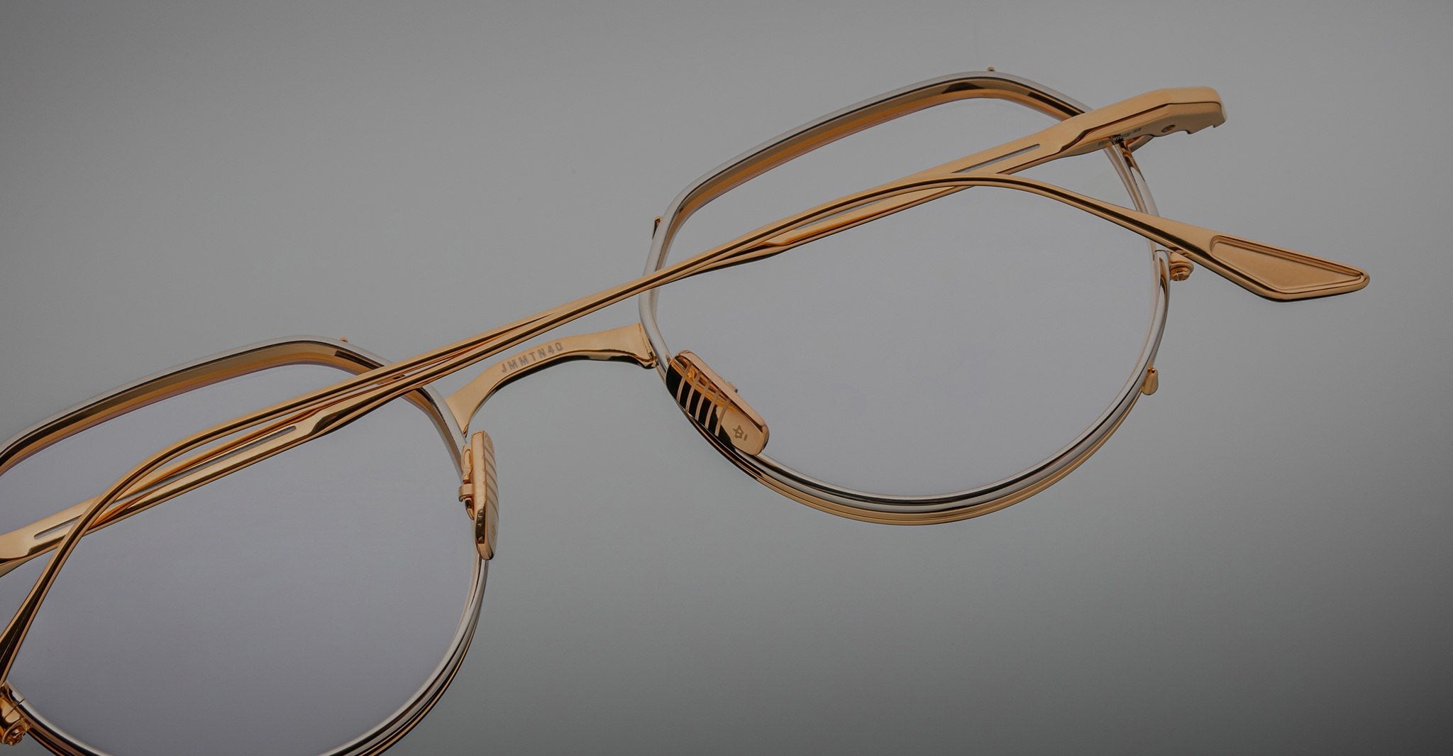 Close-up of Jacques Marie Mage Hartana gold-framed eyeglasses with clear lenses on a neutral gray background, highlighting the frame’s fine details and craftsmanship.
