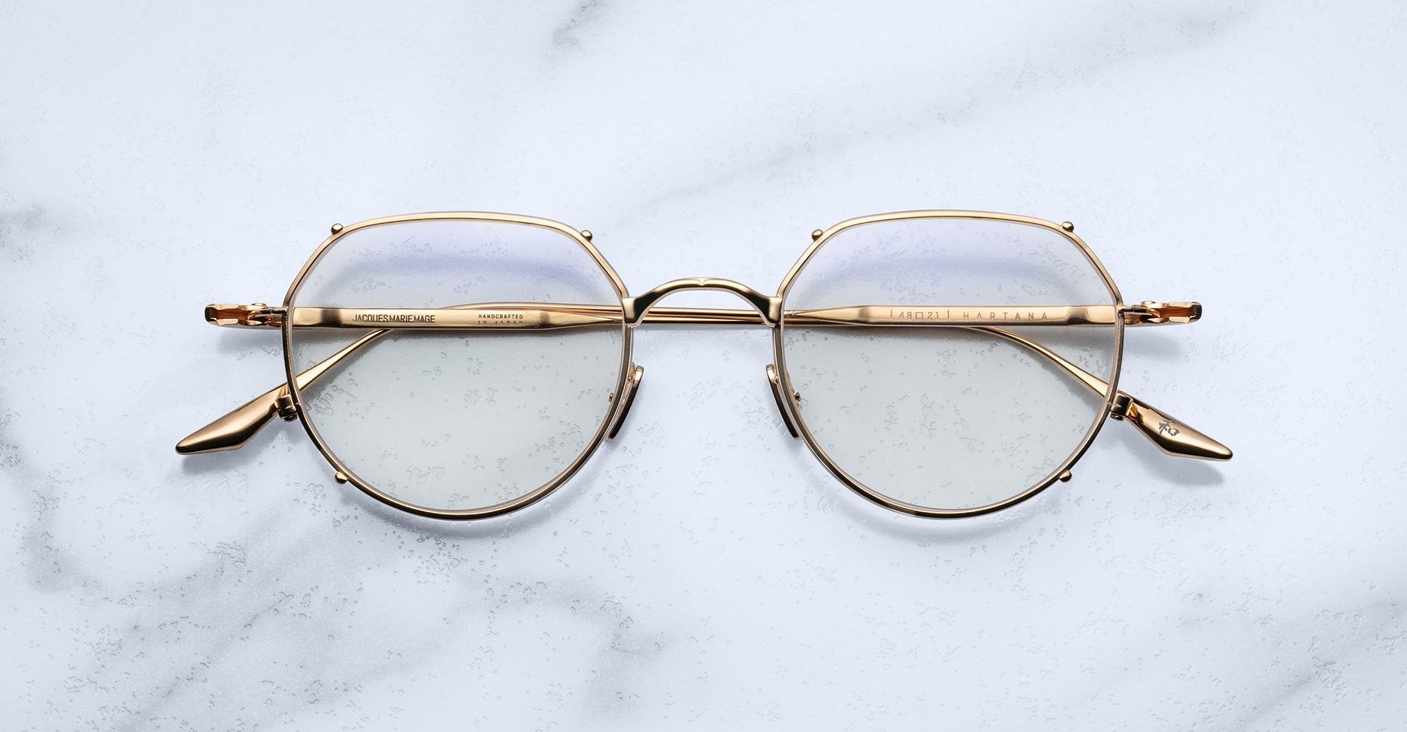 The Jacques Marie Mage Hartana gold metal round eyeglasses with clear lenses rest on a white marble surface, featuring subtly engraved arms and visible nose pads.